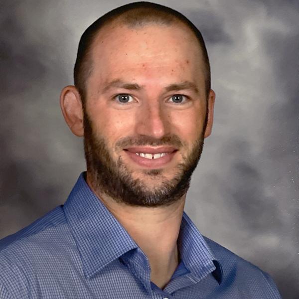 Professional portrait of a man smiling, wearing a blue polo shirt, against a dark gray background.