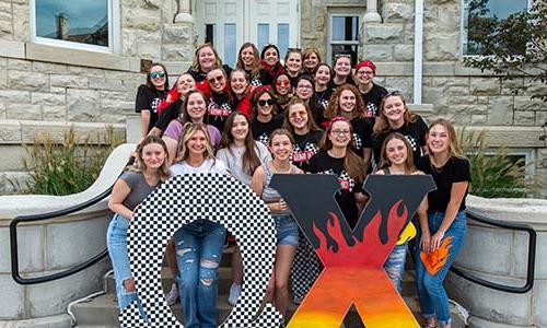 A group of smiling young women posing for a photo together, some holding symbols or letters, suggesting they might be part of a sorority or student organization.